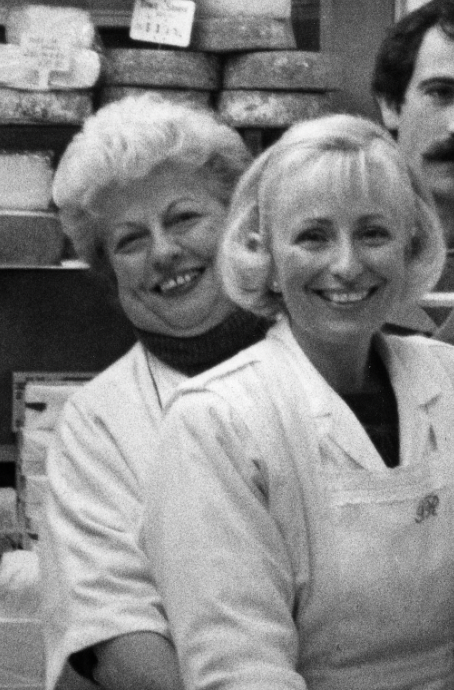 La Mère Richard, en noir et blanc, dans sa fromagerie d’époque, tenant fièrement un plateau de fromages, incarnant la tradition et l’authenticité de la gastronomie lyonnaise.